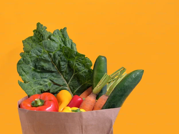 Fresh vegetables including carrots, cucumbers, and bell peppers in a brown paper bag against an orange background